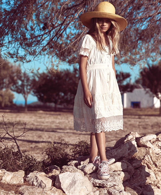 A young girl stands on a stone wall wearing a white eyelet dress, straw hat, and pink sneakers, photographed in a rural outdoor setting.