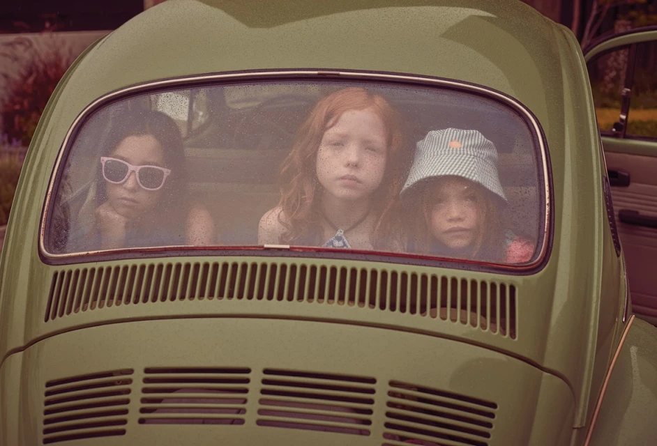 Three young children inside a vintage car, looking out the back window. A sunny day with vintage aesthetic and natural outdoor setting.