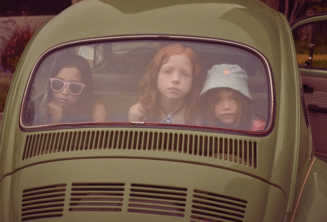 Three young children inside a vintage car, looking out the back window. A sunny day with vintage aesthetic and natural outdoor setting.