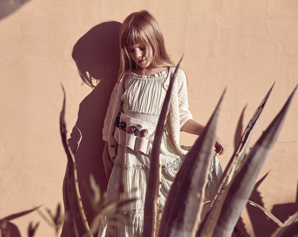 A young girl with long hair stands in front of a textured peach wall wearing a pastel dress and a tool belt with objects.