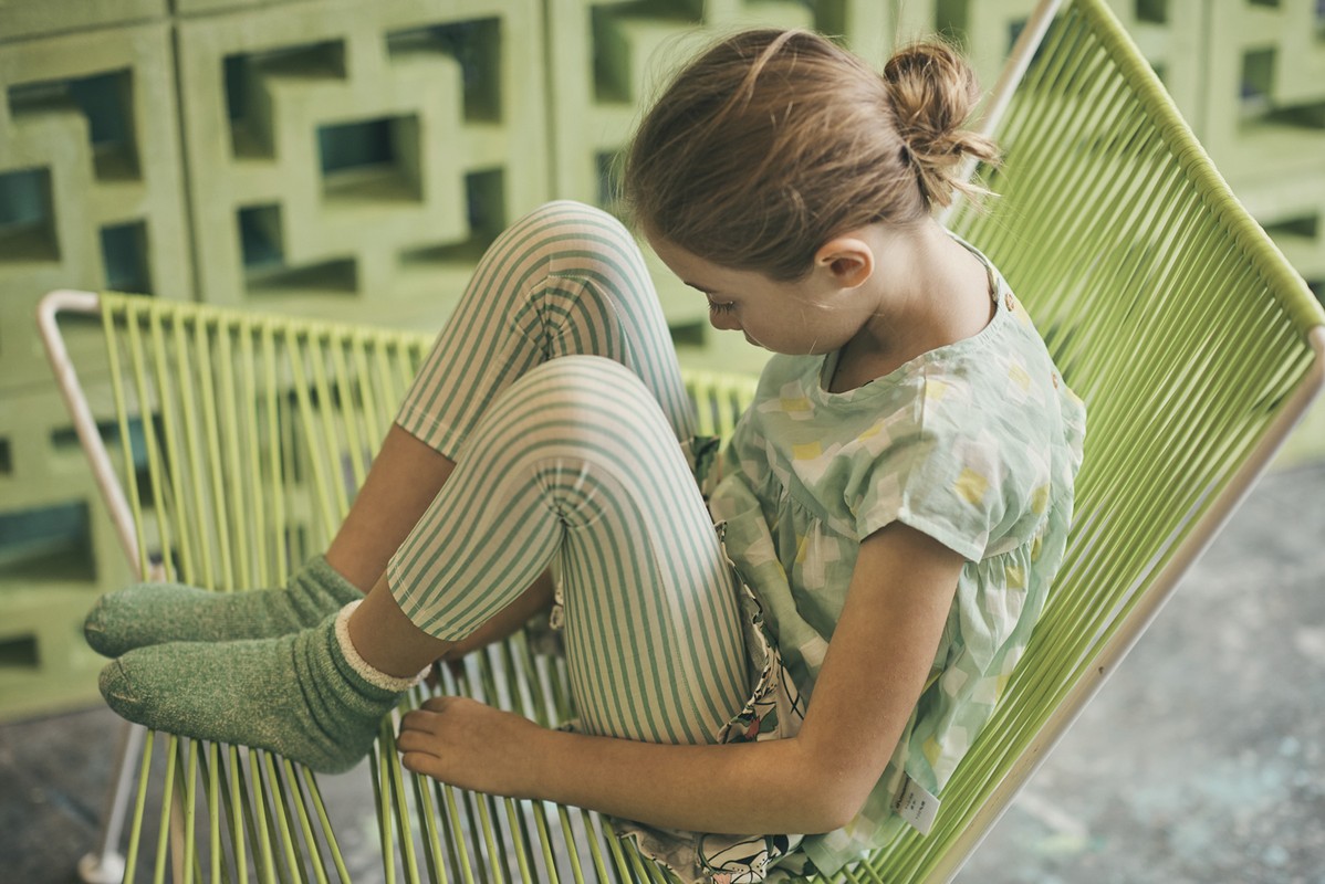 Young girl with light-brown hair sits in a lime-green rope chair wearing a light-green patterned shirt and striped pants and socks.