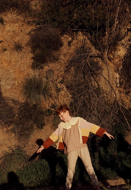 Red-haired person in beige pants, shirt, and cardigan stands outdoors near a dirt hill and dried foliage, with arms slightly outstretched.