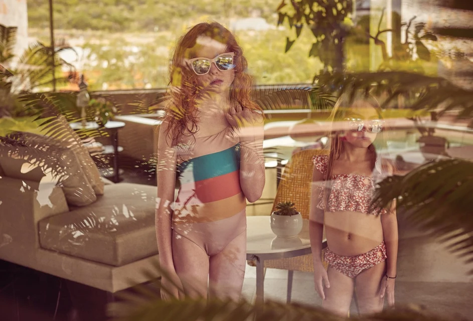 Two young girls in swimsuits and sunglasses stand outside near a pool and tropical foliage.  Bright, sun-drenched daytime scene.