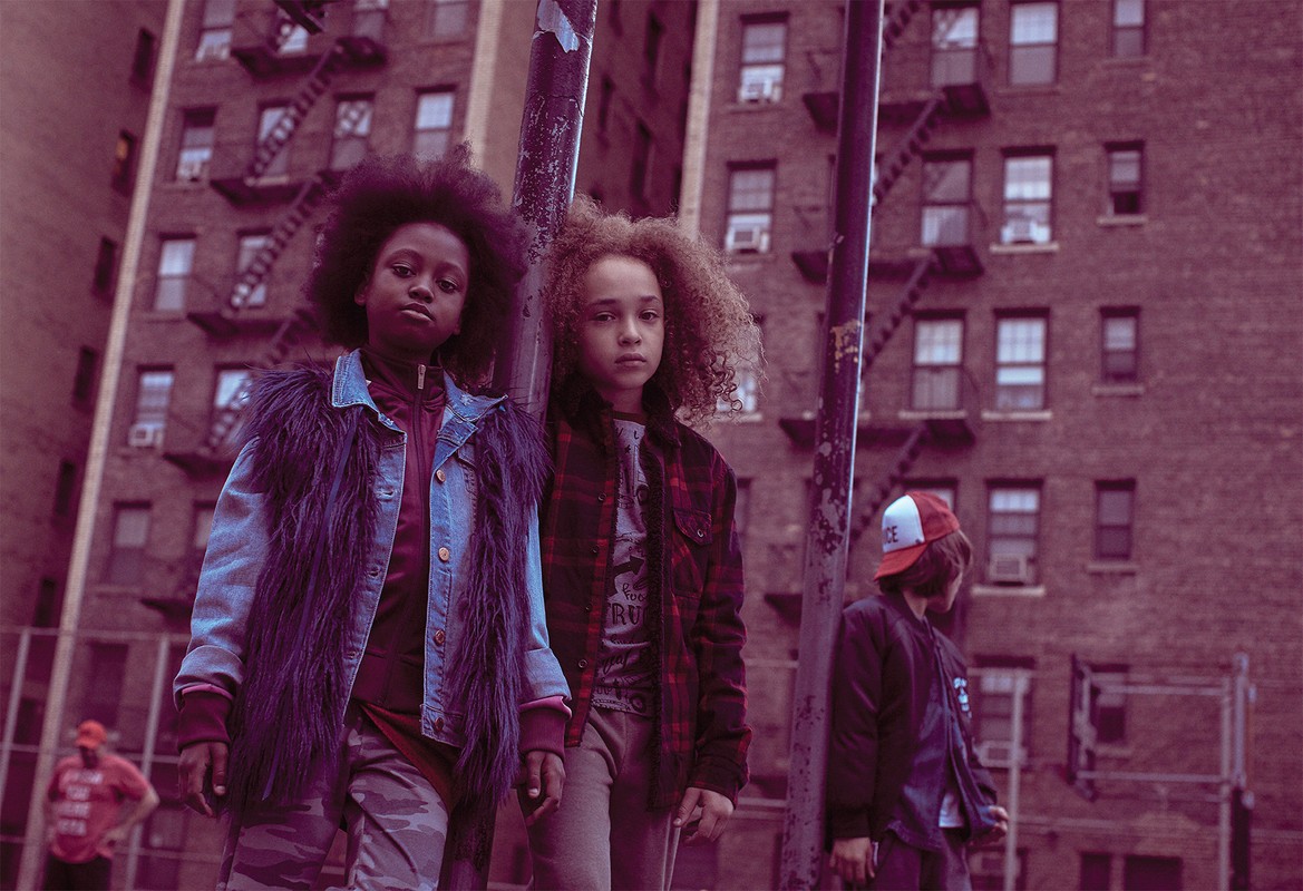 Three children stand in an urban setting, with a brick building as a backdrop. One wears a denim jacket with fur, another a plaid shirt.