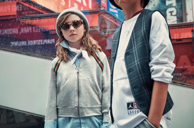 Two young children wearing fashionable clothing stand outdoors against a blurred urban backdrop with reflections and soft, diffused lighting.