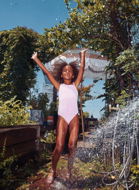 A girl in a swimsuit stands with arms raised outdoors under a water spray in a lush backyard, capturing a joyful summertime scene.