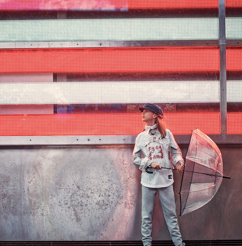Young model holding clear umbrella wearing patterned shirt and cap standing before illuminated neon sign grid
