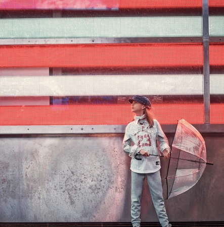 Young model holding clear umbrella wearing patterned shirt and cap standing before illuminated neon sign grid