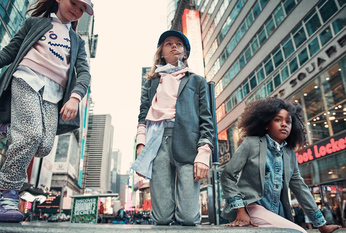 Three young girls in casual fashion pose in an urban setting with high-rise buildings and billboards, capturing a candid, modern vibe.