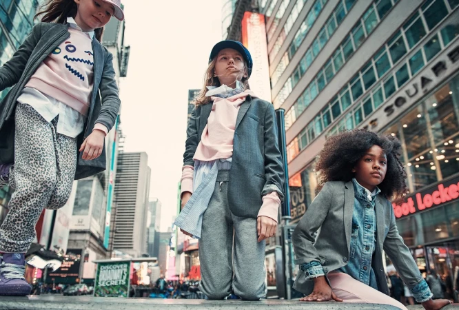 Three young girls in casual fashion pose in an urban setting with high-rise buildings and billboards, capturing a candid, modern vibe.