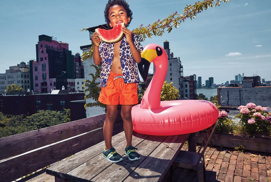 A young boy on a rooftop deck eats watermelon next to a pink flamingo pool float with a city skyline in the background on a sunny day.