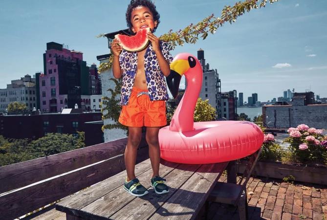 A young boy on a rooftop deck eats watermelon next to a pink flamingo pool float with a city skyline in the background on a sunny day.