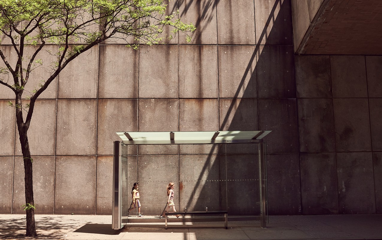 Two young girls stand inside a glass bus stop shelter on a city sidewalk next to a mature tree against a textured concrete wall.