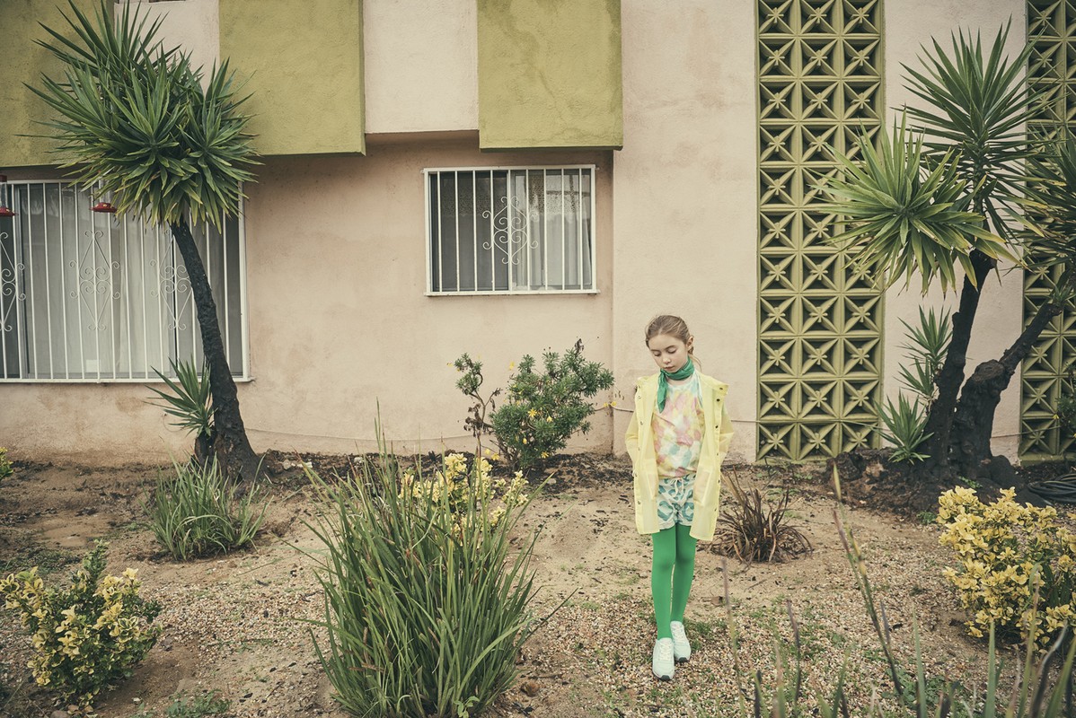 Young girl stands in a lush garden wearing a yellow jacket, green leggings, and white sneakers. She is posed in front of a peach-colored building.