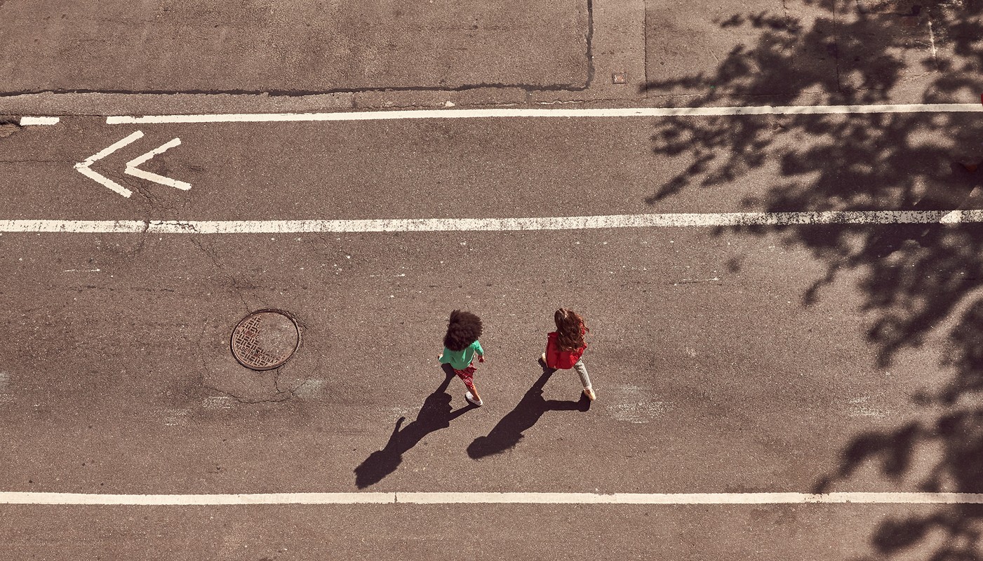 Two children walk down an asphalt street with white lines and a tree shadow, shot from an overhead angle on a sunny day.