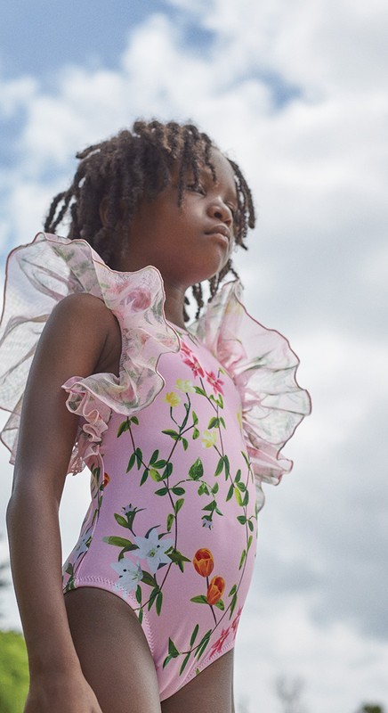 Young girl in a floral-patterned swimsuit with sheer ruffle sleeves gazes upward on a bright day with a blue sky and clouds overhead.