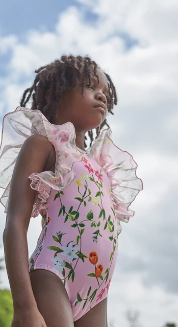 Young girl in a floral-patterned swimsuit with sheer ruffle sleeves gazes upward on a bright day with a blue sky and clouds overhead.
