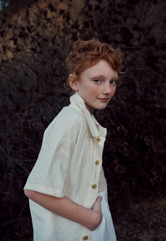 Portrait of a fair-skinned child with red hair, wearing an off-white shirt, standing against a dark, textured background. The shot is medium.