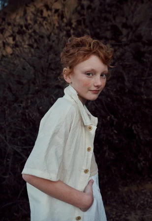 Portrait of a fair-skinned child with red hair, wearing an off-white shirt, standing against a dark, textured background. The shot is medium.