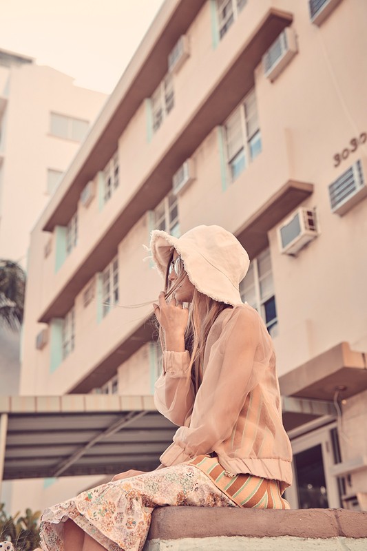 Woman in hat sits outdoors in neutral tones, wearing patterned skirt and jacket, with a building in the background on a sunny day.