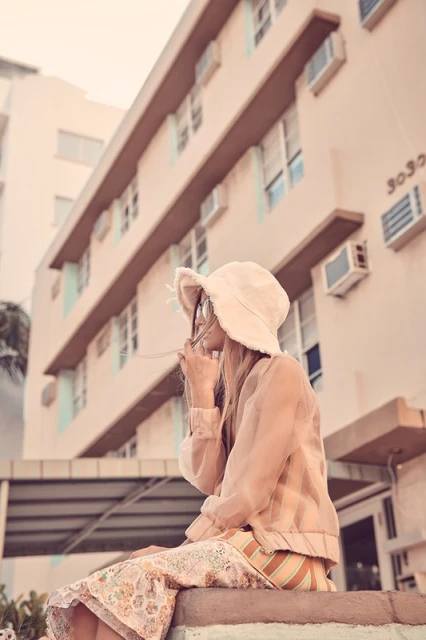 Woman in hat sits outdoors in neutral tones, wearing patterned skirt and jacket, with a building in the background on a sunny day.