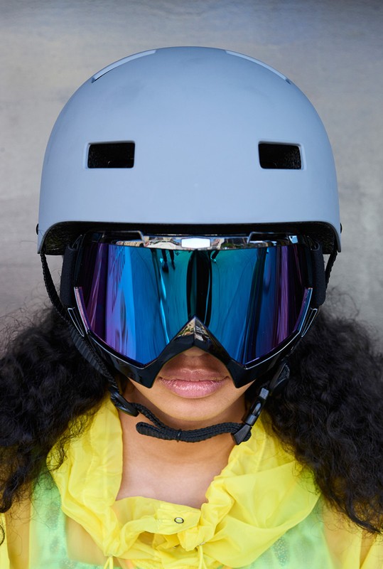 Woman wearing a grey helmet and iridescent goggles; she is wearing a yellow jacket and has curly dark hair with a neutral expression.