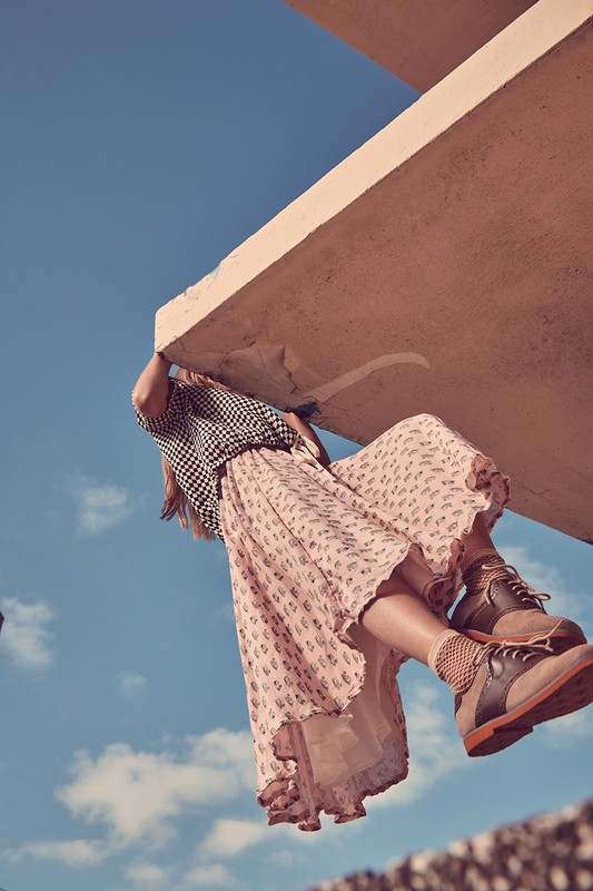 Woman in patterned blouse and skirt hanging from a concrete ledge against a blue sky in an outdoor fashion shoot.