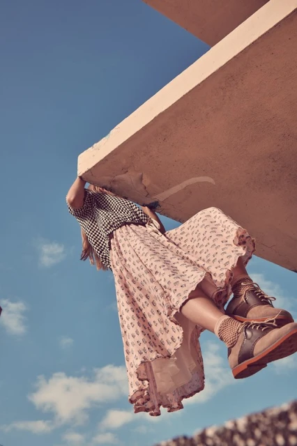 Woman in patterned blouse and skirt hanging from a concrete ledge against a blue sky in an outdoor fashion shoot.