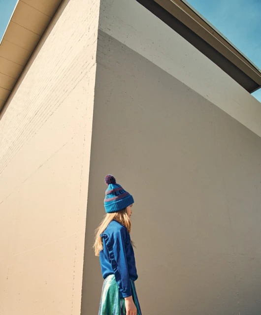 A young girl in a blue hat and matching shirt stands against a modern concrete wall outdoors on a sunny day.