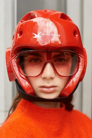 Person wearing a shiny red martial arts head guard with clear eye shield and a red turtle neck sweater indoors. Close-up studio shot.