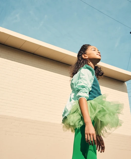 Young girl with curly hair, wearing green tutu and jacket, stands in sunlight against a cream wall and blue sky in an urban setting.