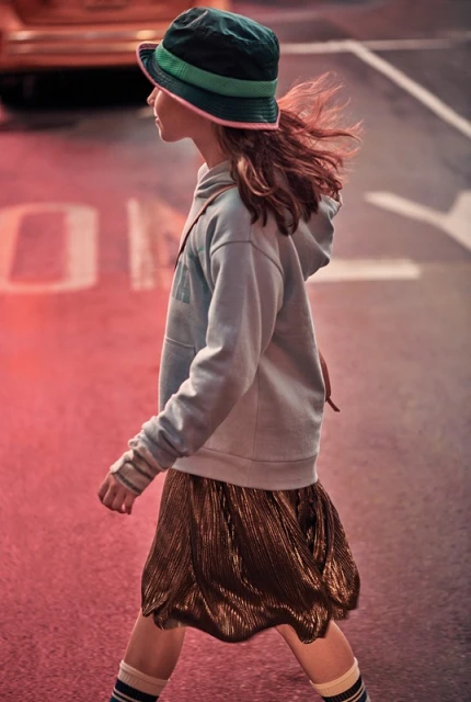 Young girl with wavy hair wearing a bucket hat, light blue hoodie, pleated skirt, and striped socks crossing a city street.