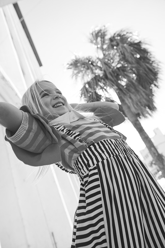 Low-angle, black and white shot of a blonde child in a striped outfit laughing, with a palm tree and bright sky in the background.