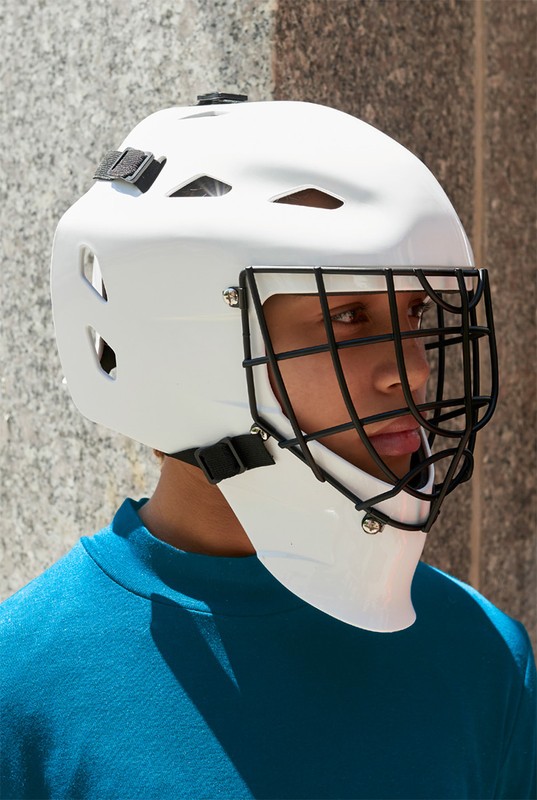 Portrait of a young adult wearing a white hockey mask and teal shirt, standing against a gray concrete wall, looking off-camera.