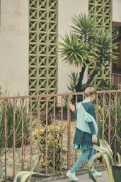 Young child walking past a metal fence wearing a dark dress with blue tights and sneakers in Los Angeles.