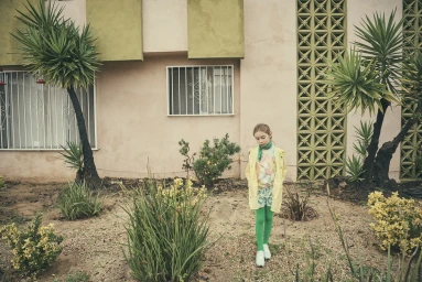 Young girl in a yellow raincoat and green tights standing outdoors near a beige building with tropical plants and decorative breeze blocks.