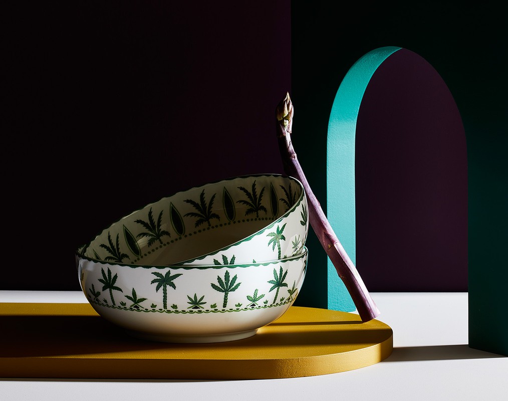 Still life of stacked patterned bowls and asparagus in front of colorful geometric backdrop with teal arch and deep purple wall.