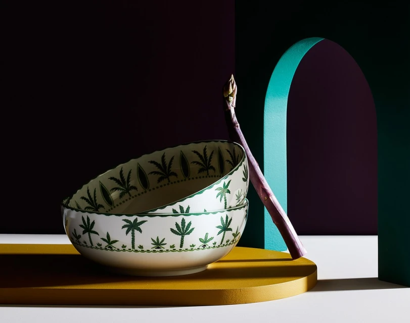 Still life of stacked patterned bowls and asparagus in front of colorful geometric backdrop with teal arch and deep purple wall.
