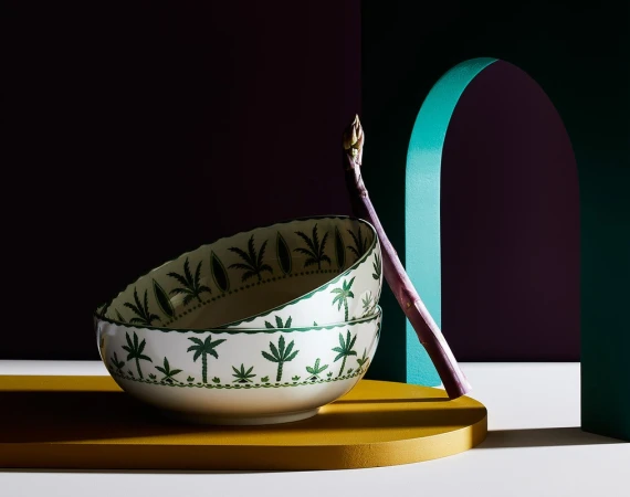 Still life of stacked patterned bowls and asparagus in front of colorful geometric backdrop with teal arch and deep purple wall.