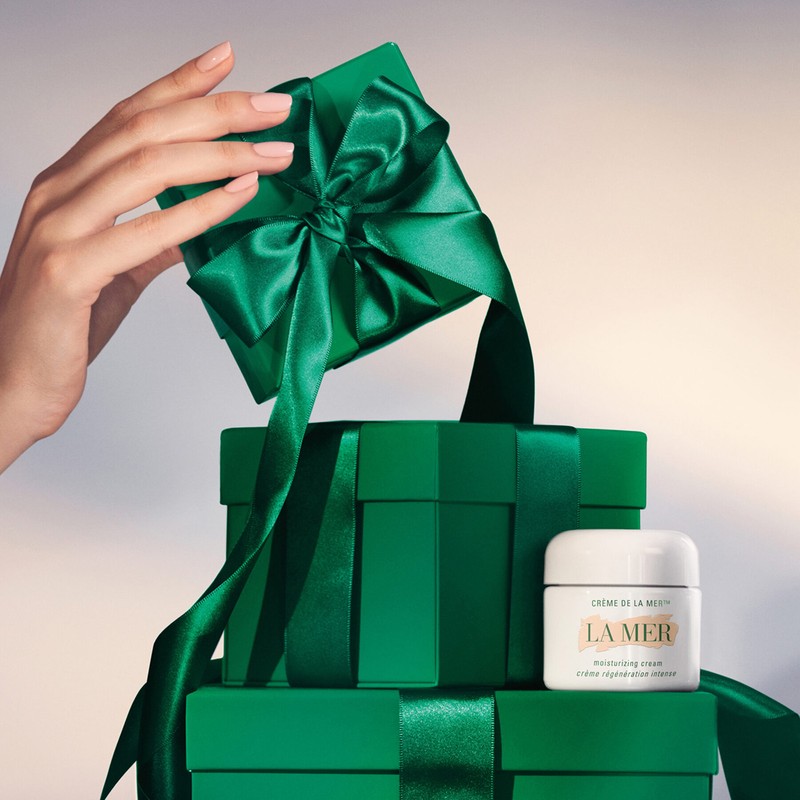 A hand opening a small green gift box tied with a shiny green ribbon, with stacked presents and a jar of La Mer cream nearby.