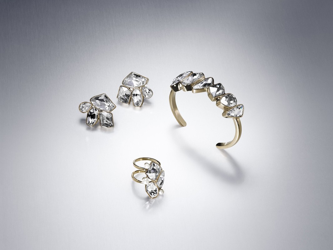 Elevated studio shot displays a ring, earrings, and cuff bracelet set with gold bands and large clear gem accents against a light grey backdrop.