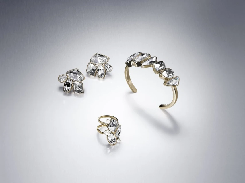 Elevated studio shot displays a ring, earrings, and cuff bracelet set with gold bands and large clear gem accents against a light grey backdrop.