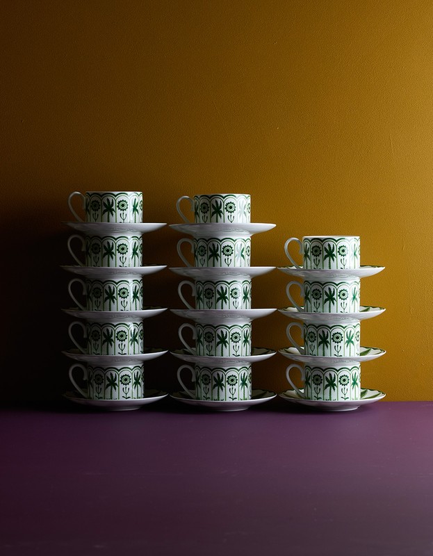 Arrangement of stacked tea cups and saucers with a palm tree pattern, set against a mustard and purple background in soft studio lighting.