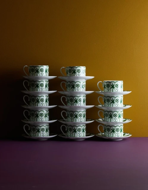 Arrangement of stacked tea cups and saucers with a palm tree pattern, set against a mustard and purple background in soft studio lighting.