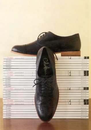 Two black leather dress shoes are stacked on a pile of magazines, one shoe standing upright to display the Cole Haan logo.