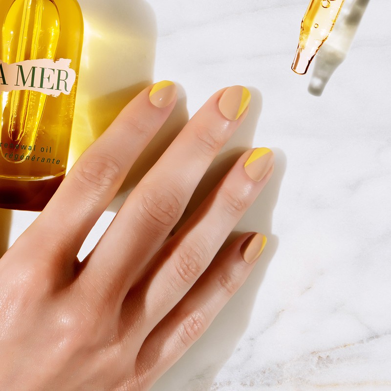 Close-up shot of a woman's hand with a two-tone yellow and beige manicure, next to La Mer skincare oil bottles and dropper against a marble surface.