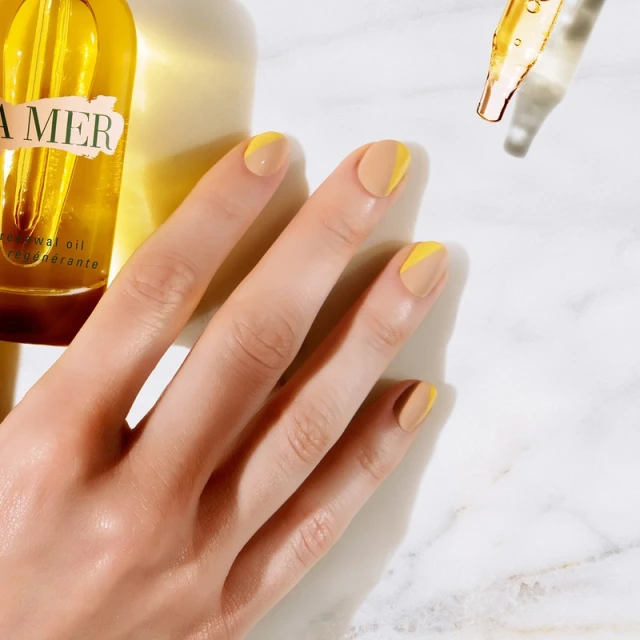 Close-up shot of a woman's hand with a two-tone yellow and beige manicure, next to La Mer skincare oil bottles and dropper against a marble surface.