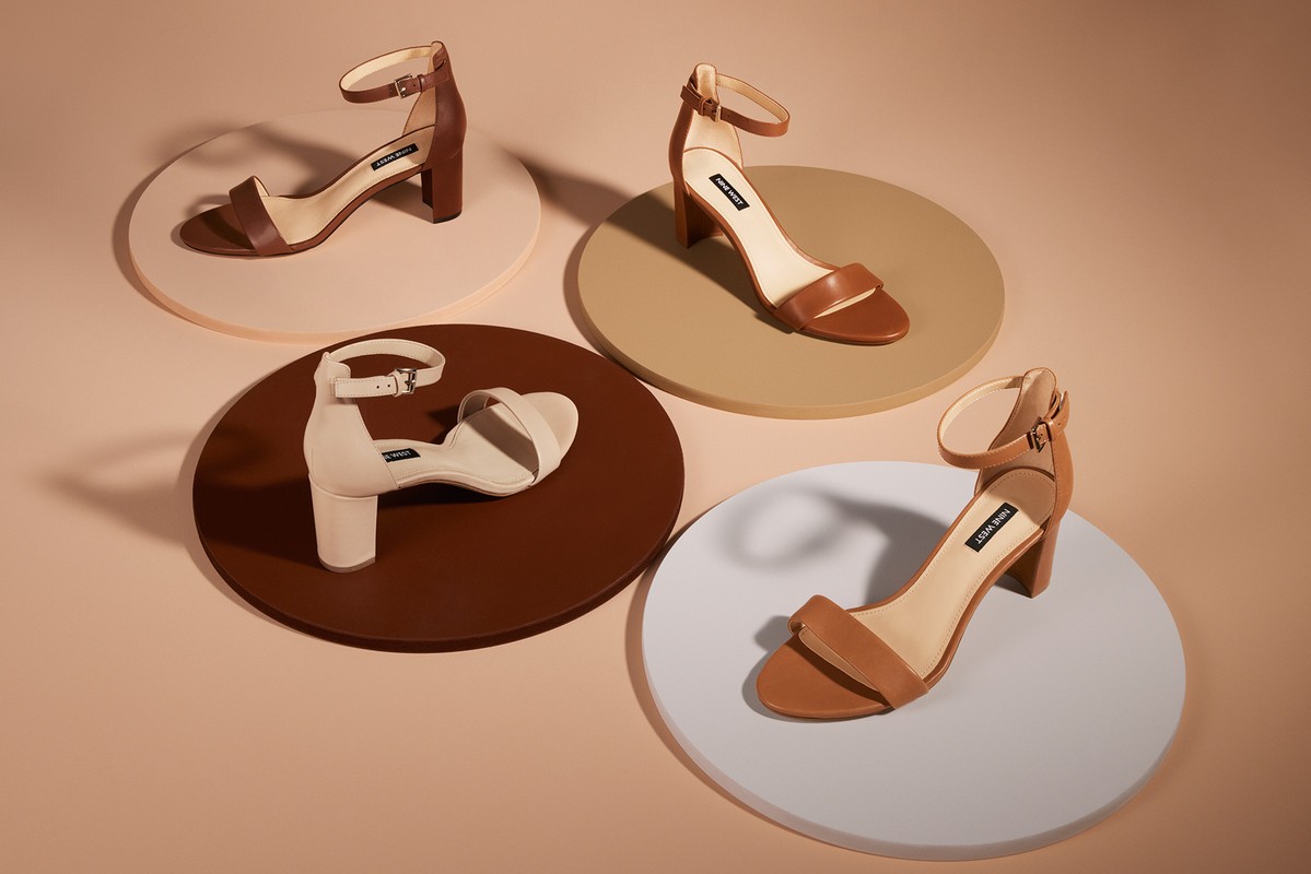 Four Nine West heeled sandals in varying colors are arranged on brown, beige, and white circular platforms against a light beige background.