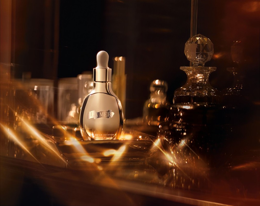 Close-up studio shot of a gold La Mer serum bottle with a dropper, set against a dark background with light flares and glass bottles.
