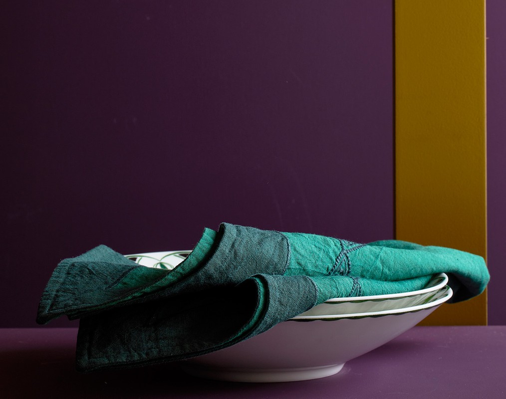 Close-up still life of a white bowl holding a folded teal linen napkin against a backdrop of purple and gold panels.
